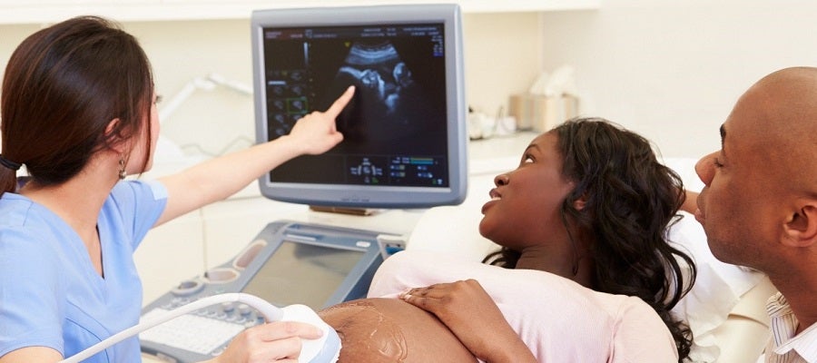 A clinician performs an ultrasound on a Black pregnant woman with her husband sitting next to her watching the image on the monitor.