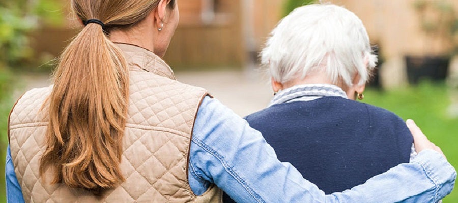 An elderly woman is escorted by a younger woman who has her arm around the shoulders of the elderly woman, viewed from behind.