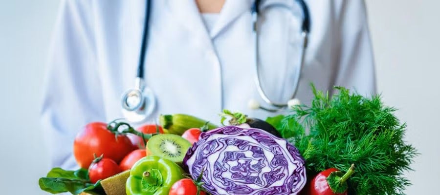 Food a medicine. A clinician in a lab coat and with a stethoscope around her neck holds a basket of assorted vegetables,